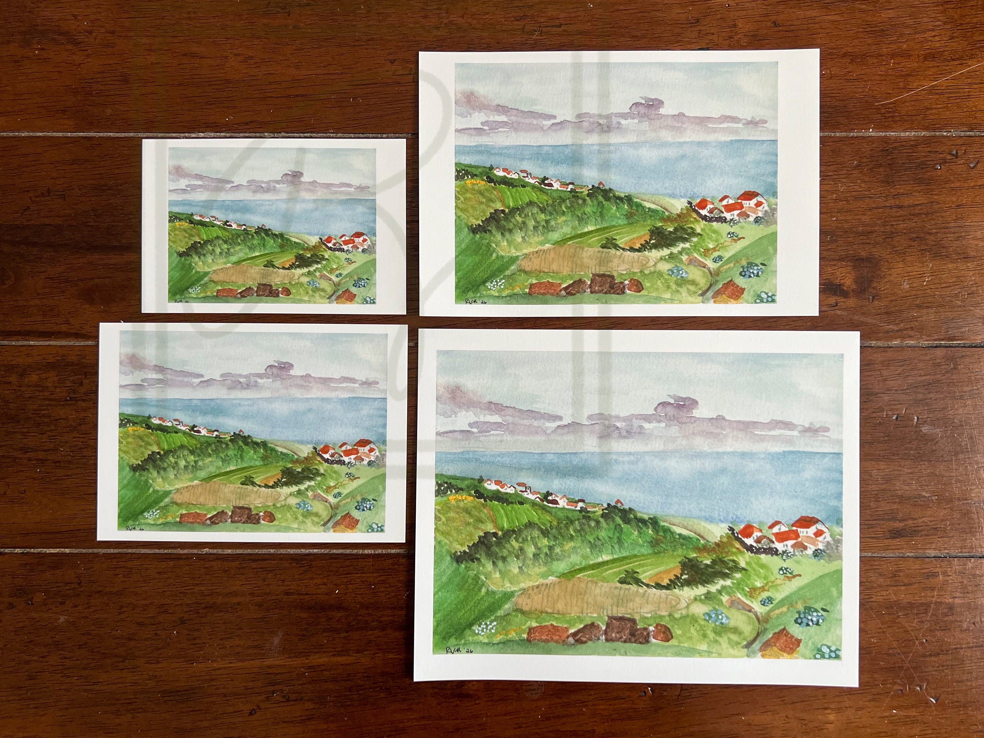 Four watercolor landscape art prints depicting green coastal hills, fields, and a blue sea under a cloudy sky, arranged on a dark wooden surface.