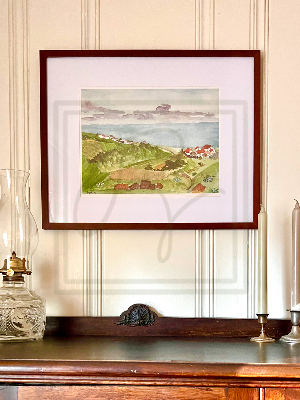 A framed watercolor landscape print of a coastal village with green hills, red-roofed houses, and a blue sea, displayed on a wooden console against a cream paneled wall.