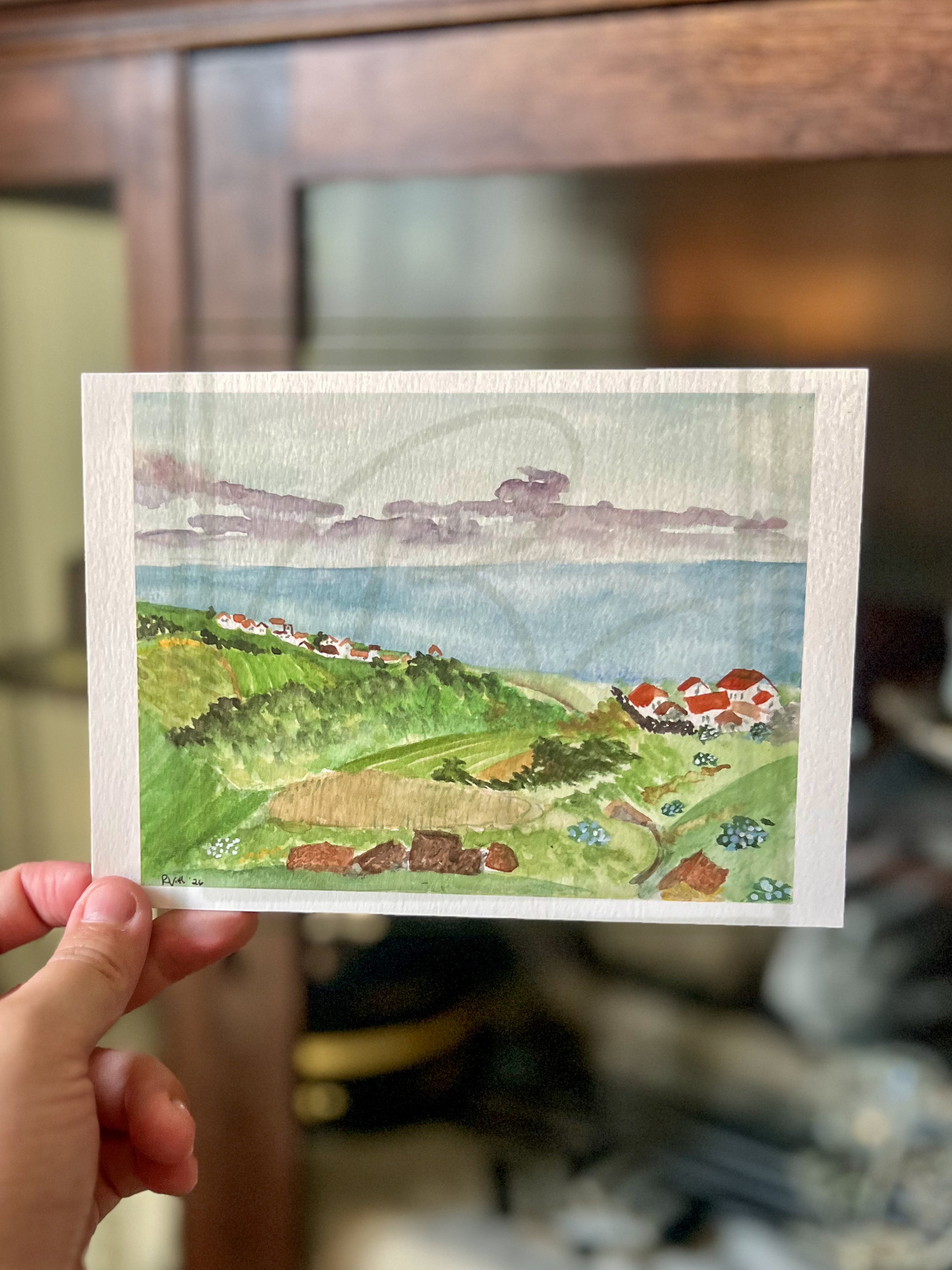 A hand holds a small watercolor landscape print showing green hills, blue water, and houses with red roofs, against a blurred indoor background with a wooden cabinet.