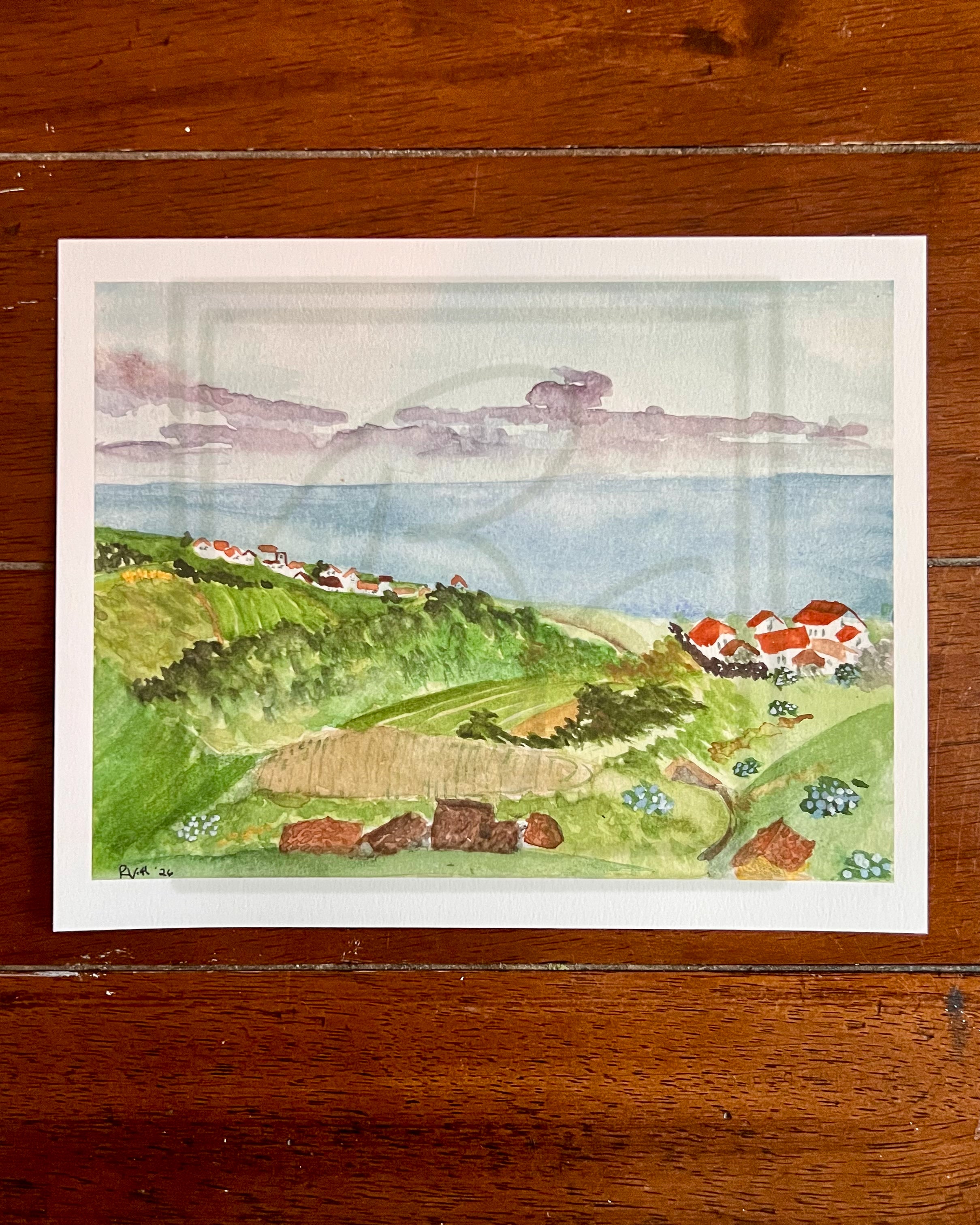 A watercolor landscape art print depicting a coastal hillside with green fields, small houses with red roofs, and a blue sea under a cloudy sky, displayed on a dark wood background.