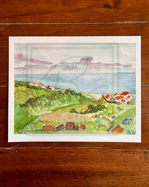 A watercolor landscape art print depicting a coastal hillside with green fields, small houses with red roofs, and a blue sea under a cloudy sky, displayed on a dark wood background.